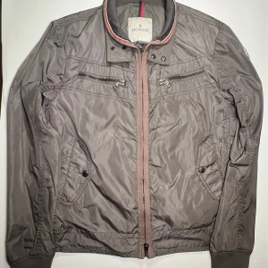 Windbreaker från Moncler - Snygg grå jacka från Moncler med dragkedja och knappar. Jackan har flera praktiska fickor med dragkedjor och en stilren design. Storlek 3 vilket motsvarar medium men passar bra ändå om du har mindre storlek 