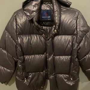 Svart dunjacka från Moncler - Säljer en snygg svart dunjacka från Moncler med huva och dragkedja. Jackan har en quiltad design och långa ärmar. Perfekt för kyliga dagar.