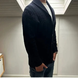 Mörkblå stickad cardigan med knappar - Säljer en mörkblå stickad kofta med knappar framtill. Koftan har ett snyggt kabelstickat mönster och långa ärmar. Perfekt för kyligare dagar.