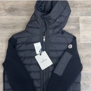 Moncler cardigan.  - Snygg svart pufferjacka från Moncler med huva och dragkedja. Jackan har långa ärmar och ribbade detaljer på sidorna. Perfekt för kyligare dagar. Kom privat för mer information 