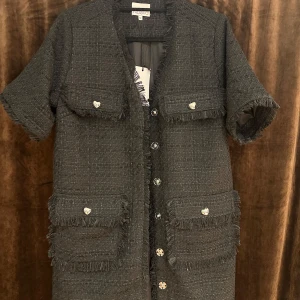 Svart kappklänning från Noella - Snygg svart klänning från Noella med korta ärmar och fransiga detaljer. Kappan har flera fickor med hjärtformade knappar och en stilren design. Perfekt för en chic look.