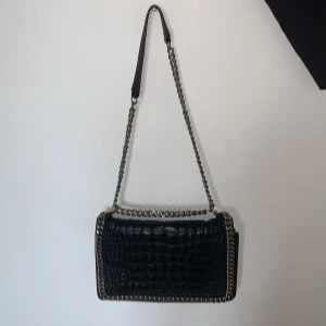 Svart axelväska från Zara Woman - Snygg svart axelväska från Zara Woman med krokodilpräglat mönster och kedjedetaljer. Väskan har en stilren design med en justerbar axelrem i kedja och läder. Perfekt för att ge din outfit en edgy touch.