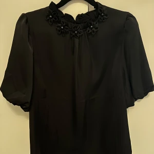 Svart blus med blommiga detaljer - Elegant svart blus i sidenmatrtial med korta ärmar och blommiga detaljer vid halsringningen. Märket är Elli White. Helt oanvänd 💕