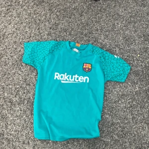 Turkos FC Barcelona fotbollströja - Säljer en turkos FC Barcelona fotbollströja med korta ärmar. Tröjan har ett unikt mönster på ärmarna och klubbens emblem på bröstet. På baksidan finns namnet 'Ter Stegen' och nummer 1 i gult. Perfekt för fans av laget!