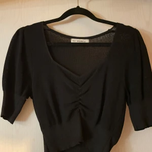 Svart topp från Divided - Snygg svart topp från Divided med v-ringning och rynkdetalj framtill. Toppen har korta ärmar och är tillverkad i ett mjukt material som ger en bekväm passform. Perfekt för en stilren look.