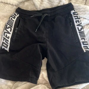 Svarta shorts från H&M med tryck för barn  - Snygga svarta shorts från H&M med coolt tryck längs sidorna. De har praktiska dragkedjor på fickorna och en bekväm snörning i midjan. Perfekta för en avslappnad stil.
