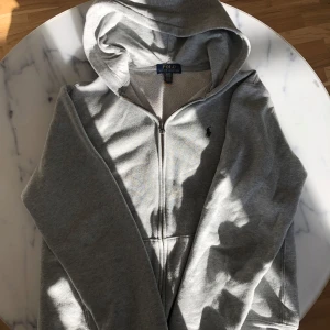 Grå hoodie från Polo Ralph Lauren - Säljer en stilren grå hoodie från Polo Ralph Lauren med dragkedja och klassisk logga på bröstet. Perfekt för en avslappnad look. Hoodien har en mjuk insida och är tillverkad i bomullsmaterial.
