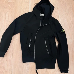 Svart jacka från Stone Island - Snygg svart jacka från Stone Island med dragkedja och huva. Storlek S, men passar även storlek M. Ikonisk jacka! 