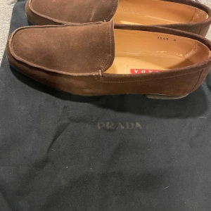 Prada Bruna mockaloafers  - Snygga bruna loafers från Prada i mjuk mocka. Skorna har en klassisk design med en diskret logotyp på innersulan och en robust sula för extra komfort. Perfekta för en stilren look. Köpta på yoox.com. Kvitto finns.