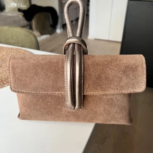 Beige mockaväska med handtag - Snygg beige väska i mocka med ett unikt handtag i metallisk ton. Väskan har en praktisk dragkedja inuti för säker förvaring. Perfekt för att ge din outfit en stilren touch.