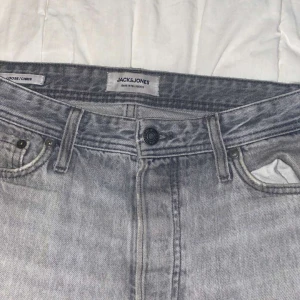 Grå jeans från Jack & Jones - Snygga grå jeans från Jack & Jones med klassisk femficksdesign och knappgylf. Perfekta för en avslappnad stil. Märkeslapp baktill i brunt läder.