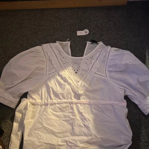 Vit blus med spetsdetaljer - Charmig vit blus med spetsdetaljer och korta puffärmar. Blusen har en v-ringad design med dekorativa broderier framtill och knytband i midjan för en justerbar passform.