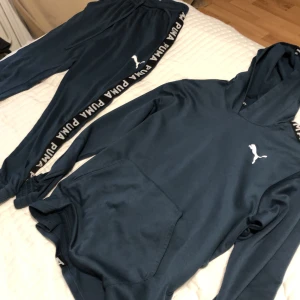 Mörkblå hoodie från Puma - Säljer en snygg mörkblå hoodie från Puma med vit logga på bröstet och text längs ärmarna. Tröjan har en praktisk magficka och en bekväm huva. Perfekt för en sportig look! Ingår också byxor det är jätte bra skick på tröjan men helt okej skick på byxorna