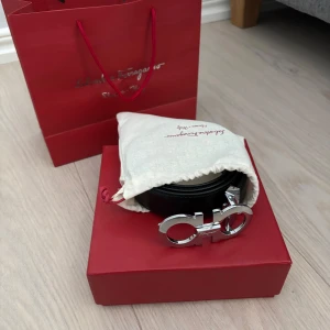 Svart bälte från Salvatore Ferragamo 110cm - Snyggt svart bälte från Salvatore Ferragamo med en stilren silverfärgad spänne (110cm). Perfekt för att ge en elegant touch till din outfit. Kommer i originalförpackning med en röd presentpåse och skyddspåse samt kvitto. Pris kan diskuteras