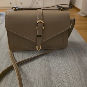Oanvänd beige axelväska med gulddetaljer - Snygg beige axelväska med justerbar axelrem och stiliga gulddetaljer. Väskan har ett elegant spänne framtill och en praktisk knappstängning.