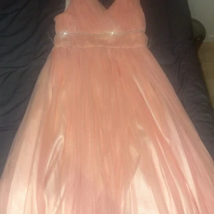 Persikofärgad klänning från Civil Girls - Elegant persikofärgad långklänning från Civil Girls med v-ringning och glittrande detaljer i midjan. Perfekt för speciella tillfällen med sin flytande design och ärmlösa stil.