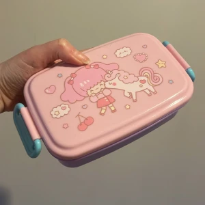 Rosa och lila matlåda med enhörningsmotiv - Söt matlåda i rosa och lila med ett gulligt motiv av en flicka och en enhörning. Lådan har ett praktiskt handtag i turkos och är perfekt för att ta med lunch eller snacks. Den är lätt att öppna och stänga, vilket gör den idealisk för både barn och vuxna. Foto 2 visar en liten defekt, annars i mycket fint skick 