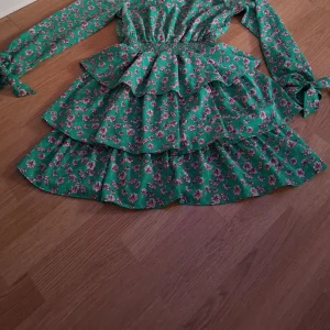 Grön blommig klänning med volanger - Charmig grön klänning med rosa blommor och volanger. Den har långa ärmar med knytband och en smickrande v-ringning. Perfekt för en somrig look!