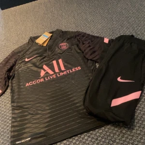 Svart och rosa PSG fotbollströja från Nike - Snygg svart PSG fotbollströja med rosa detaljer från Nike. Tröjan har korta ärmar och ett diskret randigt mönster. Den har PSG-loggan och texten 'Accor Live Limitless' i rosa framtill. Perfekt för fotbollsfans som vill ha en stilren look.