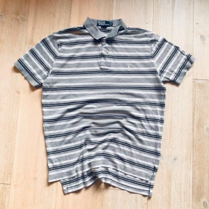 Randig pikétröja från Ralph Lauren - Säljer en klassisk grå och vit randig pikétröja från Ralph Lauren. Tröjan har korta ärmar och en broderad logga på bröstet. Tillverkad i 100% bomull för en bekväm passform. Perfekt för en stilren look. Nästan aldrig använd och inga skavanker eller defekter!