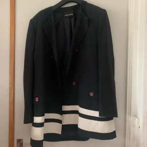 Svart och vit kavaj från Guy Laroche - Elegant svart kavaj från Guy Laroche med vita ränder längst ner. Kavajen har långa ärmar och röda knappar som ger en snygg kontrast. Perfekt för en stilren look.