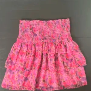 Rosa blommig kjol med volang - Snygg rosa kjol med blommigt mönster och volangdetaljer. Perfekt för en färgglad look. Kjolen har en smockad midja för en bekväm passform.