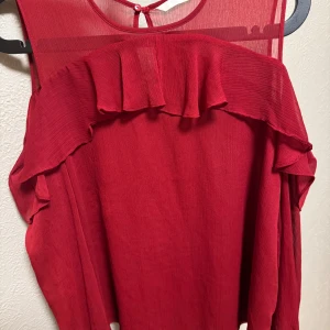 Röd ärmlös blus med volangdetaljer - Snygg röd blus med volangdetaljer och transparenta partier vid axlarna. Den är ärmlös och har en luftig passform, perfekt för varmare dagar. Klänningen har en rund halsringning och knäppning i nacken.