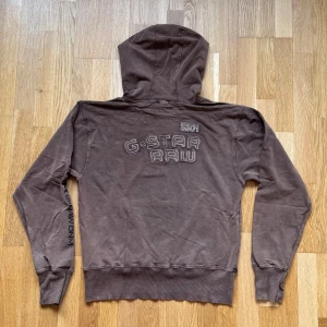 G-Star Raw Brun Hoodie - Brun Hoodie med broderat tryck både på fram och baksida från märket G-Star Raw.  Storlek: XL (Sitter liten), Armhåla till Armhåla: 52 cm, Längd: 60 cm, Axel till Armöppning: 62 cm. Material: 100% Cotton