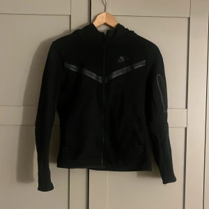 Svart hoodie från Nike - Säljer en svart hoodie från Nike med dragkedja och huva. Den har en diskret logga på bröstet och en stilren design med en mörk randdetalj över bröstet. Perfekt för en sportig look eller avslappnade dagar. Passar mig som har xs 