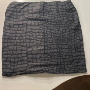 Grå kjol med krokodilmönster - Snygg grå kjol med ett coolt krokodilmönster. Perfekt för att ge din outfit en unik touch. Kjolen har en kort längd och en normal passform.