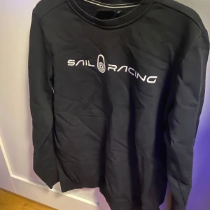 Svart tröja från Sail Racing - Skitsnygg tröja från Sail Racing i storlek 176. Jättebra skick annars men lite smått sliten text som ni kan se på andra bilden. Skulle det vara något annat så är det bara o höra av sig👍