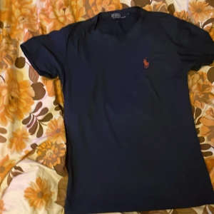 Mörkblå t-shirt från Polo Ralph Lauren - Säljer en stilren mörkblå t-shirt från Polo Ralph Lauren med en röd broderad logotyp på bröstet. T-shirten har en klassisk passform och är tillverkad i mjuk bomull. Perfekt för en avslappnad look.