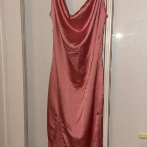 Rosa sidenklänning - Elegant rosa klänning i siden med en vacker draperad halslinje,med en slits vid benet. Klänningen har en smickrande passform och är perfekt för speciella tillfällen. Den är ärmlös och har en mjuk, glansig yta som ger en lyxig känsla.