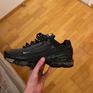 Svarta Nike sneakers - Snygga svarta Nike sneakers med en futuristisk design och synlig luftdämpning i sulan. Skorna har en liten vit Nike-logga på sidan och svarta snören. Perfekta för en sportig look.