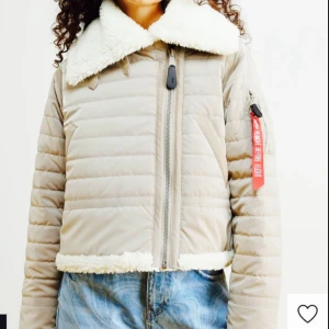 Beige pufferjacka med fuskpäls - Snygg beige pufferjacka från Alpha Industries med fuskpälsdetaljer på krage och nederkant. Jackan har en dragkedja framtill och en ikonisk röd tagg på ärmen. Perfekt för kyliga dagar med sin quiltade design. Jag köpte jackan för 2500kr men säljer den för mycket rimligt pris. Pris kan diskuteras💕