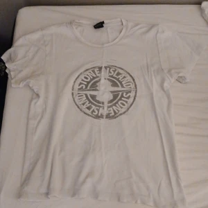 Vit t-shirt från Stone Island - Säljer en vit t-shirt från Stone Island med det ikoniska logotyptrycket på framsidan. T-shirten har en rund hals och korta ärmar. Perfekt för en avslappnad stil. Hör av er vid minsta fundering!
