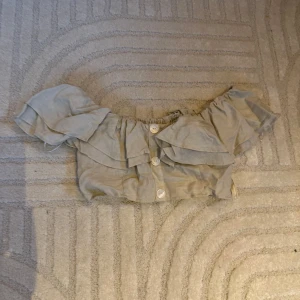 Beige offshoulder blus från Zara - Säljer en söt beige offshoulder blus från Zara med volanger och knappar framtill. Perfekt för en somrig look. Blusen har en luftig passform och är tillverkad i ett lätt material.