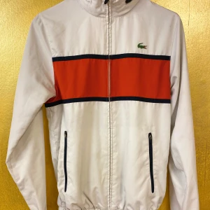 Vit vintage windbreaker från Lacoste - Klassisk, stilren vit vindjacka från Lacoste. Jackan har dragkedja och huva. I kragen. Perfekt för sportiga dagar eller en casual look. Mycket bra skick, obetydligt använd.