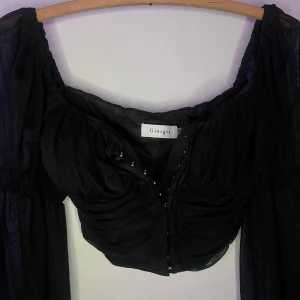 Svart offshoulder topp från Giorgia - Snygg svart offshoulder topp från Giorgia med långa ärmar i ett transparent material. Toppen har en åtsittande passform med rynkade detaljer framtill och en diskret knäppning. Perfekt för en stilren look.