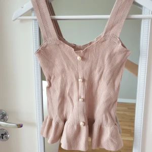 Rosa ribbad topp med volang - Söt rosa/beige ribbad topp med breda axelband och dekorativa pärlknappar framtill. Toppen har en volangdetalj vid nederkanten som ger en feminin touch.