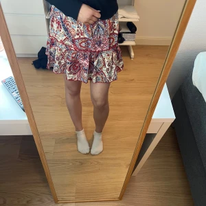 Mönstrad kortkjol med volang - Säljer en färgglad kortkjol med ett livligt paisleymönster i rött, blått och vitt. Kjolen har volangdetaljer som ger en lekfull touch. Perfekt för att liva upp garderoben!