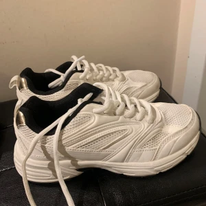 Vita sneakers - Snygga vita sneakers med gulddetaljer på hälen och svart insida. Skorna har en sportig design med snörning och en bekväm sula. Perfekta för en stilren look.