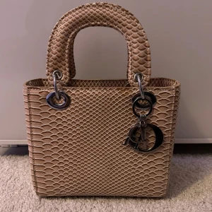 Beige handväska med ormskinnsmönster - Snygg beige handväska med ormskinnsmönster och metallringar. 