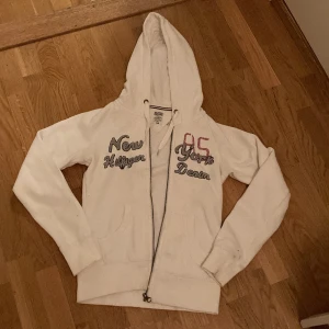 Vit hoodie från Hilfiger Denim - Säljer en vit hoodie från Hilfiger Denim med texten 'New York Hilfiger Denim' och siffran '85' på bröstet. Hoodien har dragkedja och justerbar huva med snören. Jättesnygg men har tyvärr blivit för liten. Pris kan förhandlas!