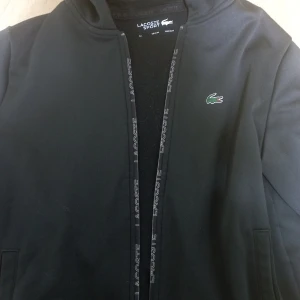 Svart jacka från Lacoste - Snygg svart jacka från Lacoste med dragkedja och det klassiska krokodilmärket på bröstet. Perfekt för en sportig look. Jackan har långa ärmar och en stilren design.