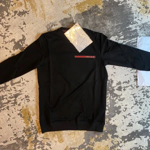 Prada tröja - Säljer en  svart tröja från Prada. Den har en enkel design med Pradas logga på bröstet och ärmen. Perfekt för både vardag och festliga tillfällen. Tröjan är i nytt skick och kommer med originaltaggar. 