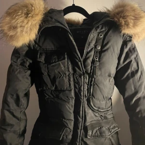 Svart parkajacka med pälsdetaljer - Snygg svart parkajacka från Hollies med en stor, fluffig pälsdetalj på huvan. Jackan har dragkedja och knappar framtill samt flera praktiska fickor. Perfekt för kalla dagar.