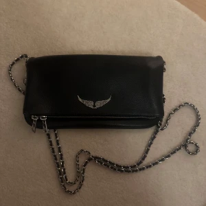 Svart axelväska med kedja - Snygg svart axelväska i skinn med en cool kedjerem och dragkedja. Väskan har en stilren design med en silverfärgad detalj framtill. Perfekt för att ge din outfit en edgy touch.