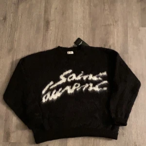 Yves Saint Laurent tröja - Säljer en snygg svart tröja från Saint Laurent med vit text på framsidan. Tröjan har långa ärmar och en rund halsringning. Perfekt för en stilren look.