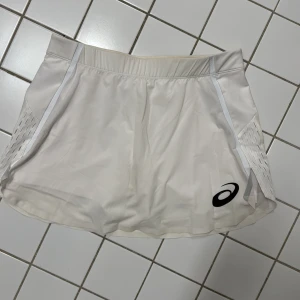 Vit kjol från Asics - Säljer en vit kjol från Asics, perfekt för sportiga aktiviteter. Kjolen har en elastisk midja och en diskret logga på framsidan. Den är designad för att ge en bekväm passform och har stilrena detaljer längs sidorna.